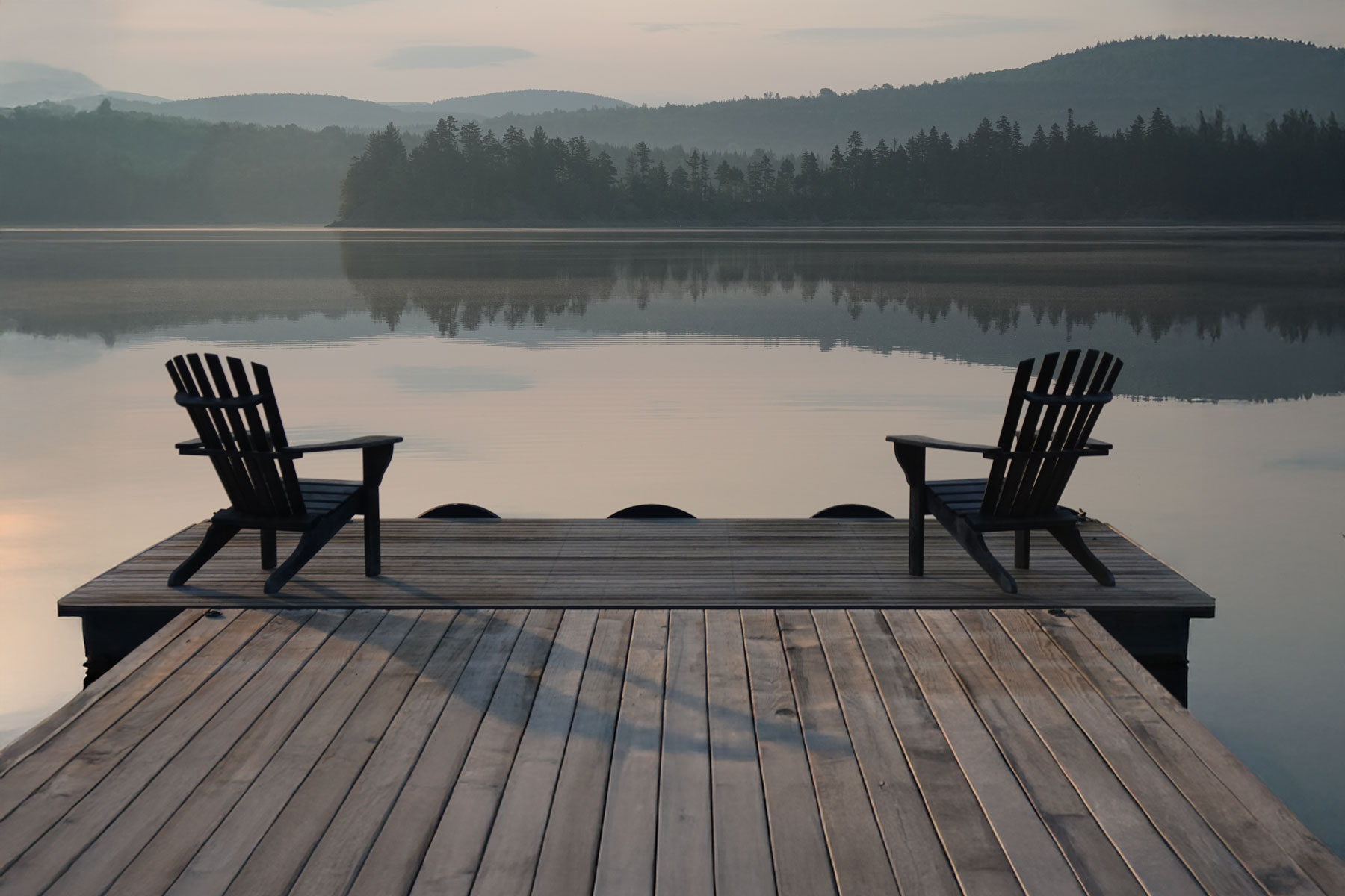 Two empty chairs facing mountains and calm lake from a quiet dock