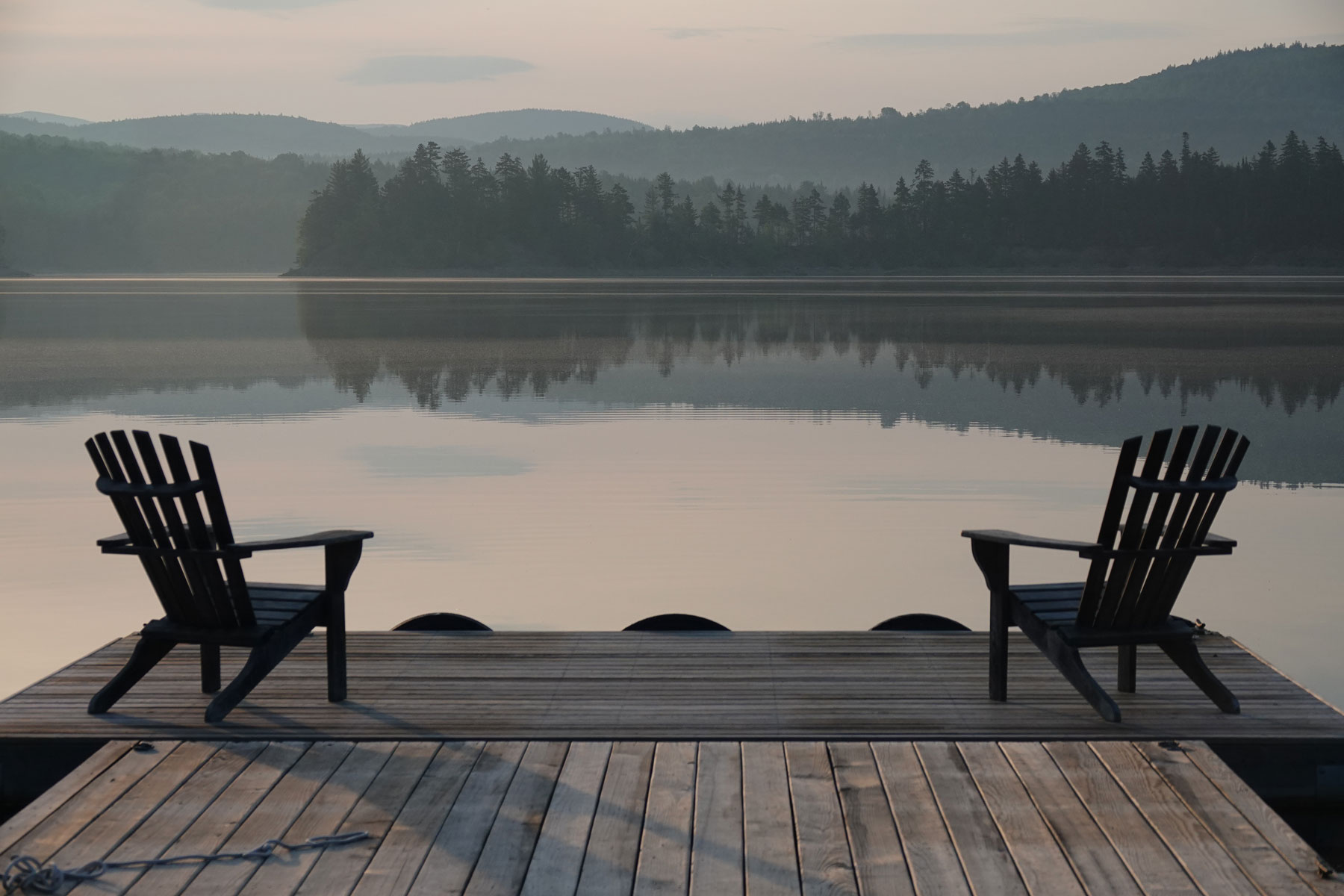 Two empty chairs facing mountains and calm lake from a quiet dock