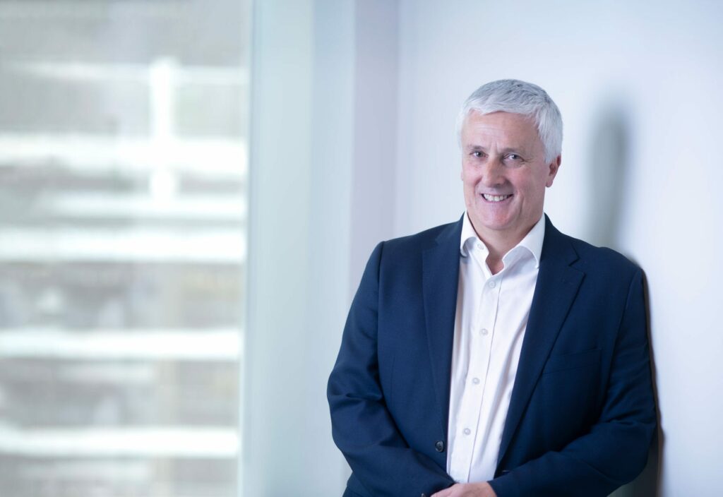 Smiling man with short grey hair in a dark suit and white shirt stands in a bright office next to a window with blurred cityscape in the background.