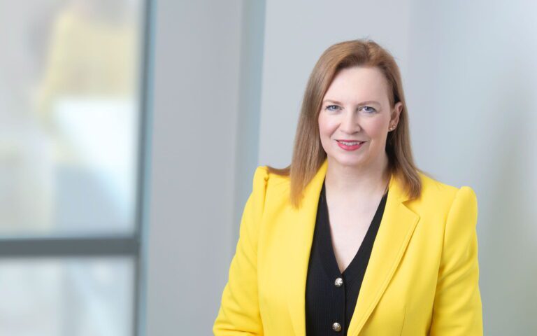 Anne Todd, a woman with straight, light brown hair, wears a bright yellow blazer over a black top, smiling whilst standing indoors near large windows with soft natural light.