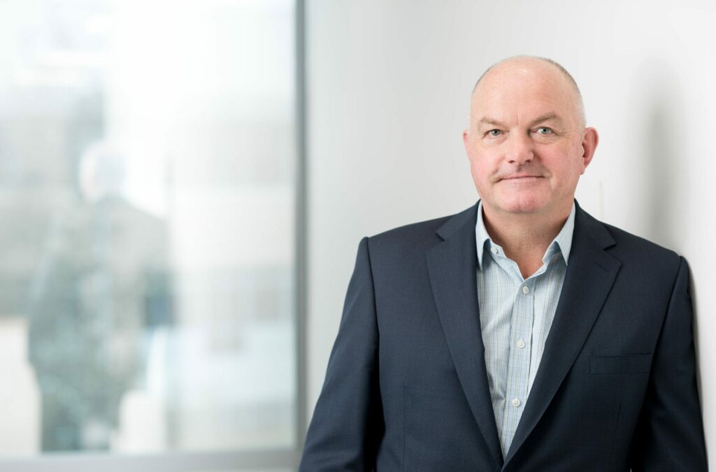 Richard Cobb, a middle-aged man in a navy suit and light blue shirt, stands indoors, leaning against a white wall with a window in the background.