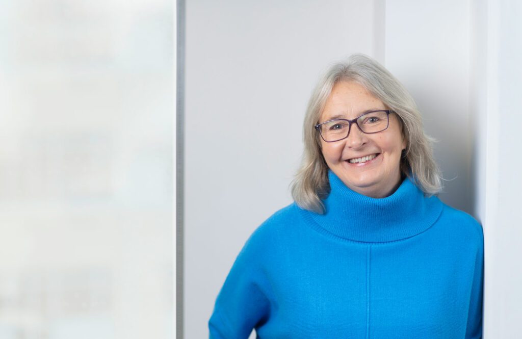 Samantha Billingham, with grey hair and glasses, wearing a bright blue polo neck jumper, stands indoors near a window with a blurred background.