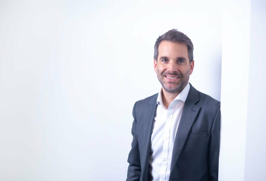 Tom Torkar - a man in a dark suit and white shirt stands smiling against a white wall with a neutral, bright background.