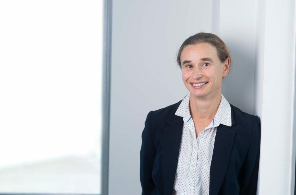 Vivienne Williams, dressed in a navy blazer and white polka dot shirt, smiles whilst leaning against a white wall in a bright office setting.