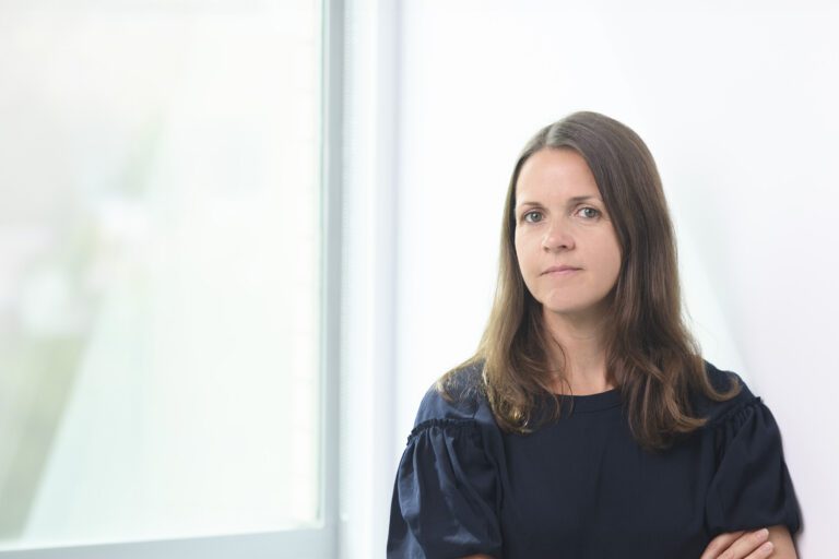 Alice Daniels with long brown hair, wearing a dark blue shirt and standing near a window.