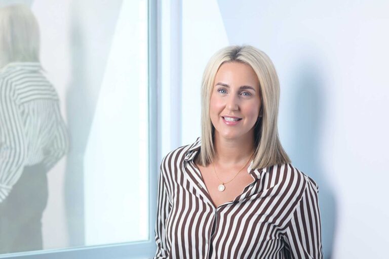 Naomi Morris, A woman with straight blonde hair wears a brown-and-white striped shirt and gold necklace, smiling whilst standing in front of a window with her reflection visible.