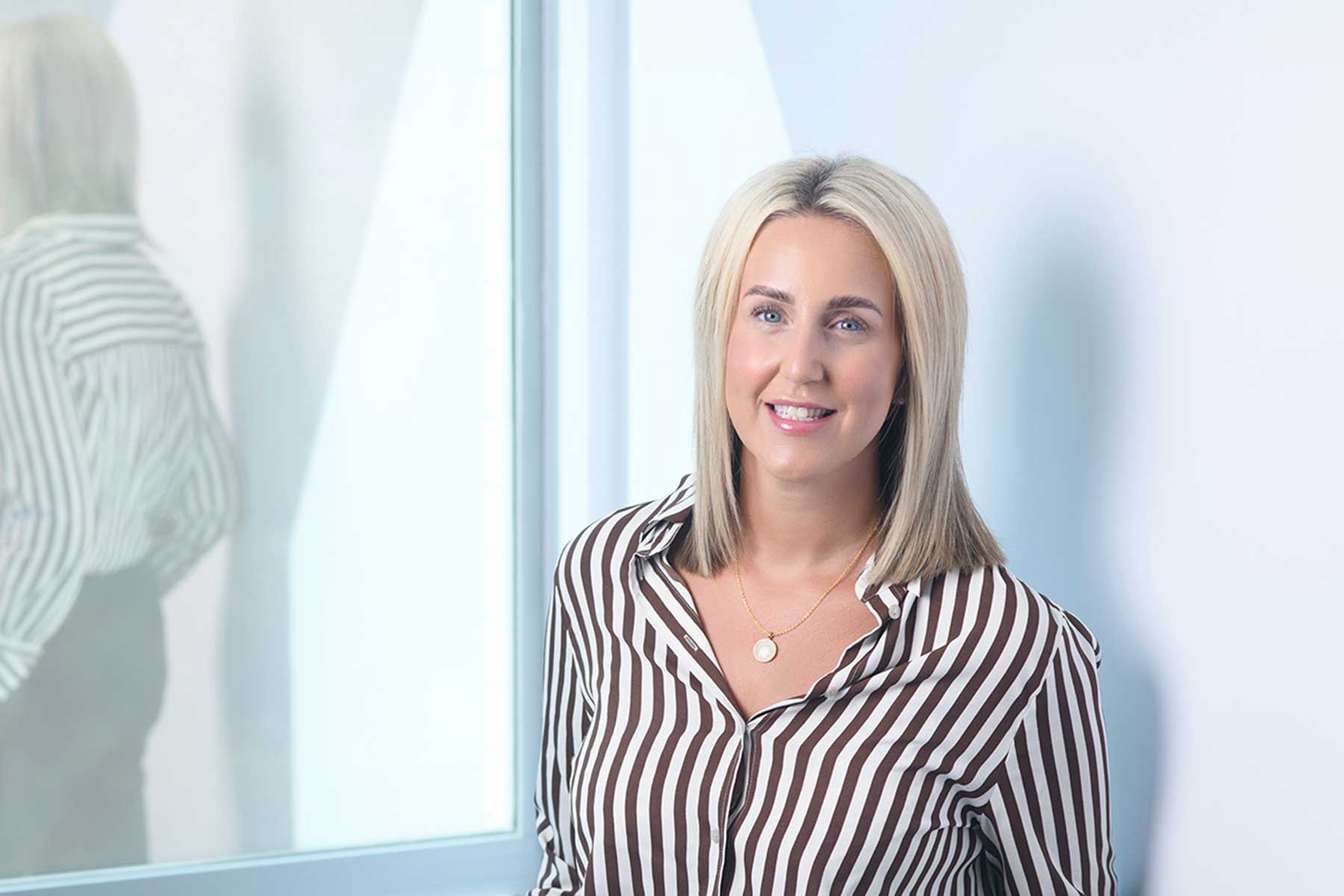 Naomi Morris, A woman with straight blonde hair wears a brown-and-white striped shirt and gold necklace, smiling whilst standing in front of a window with her reflection visible.