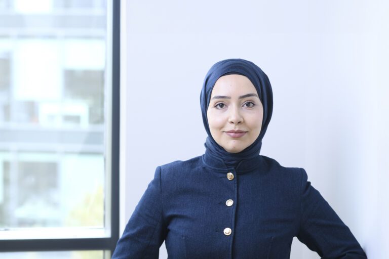 A woman, Sena Guvercin, wearing a navy blue hijab and matching buttoned top stands indoors near a window, looking confidently at the camera.