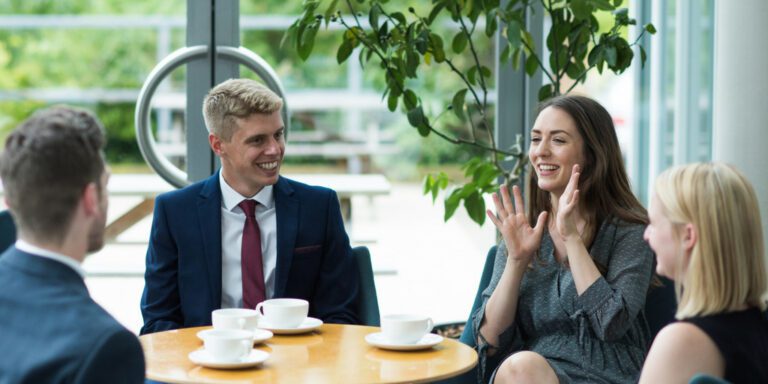 Four people in business attire sit around a table with white cups, conversing and smiling. A woman gestures animatedly with her hands. There’s a large window and green plants in the background.