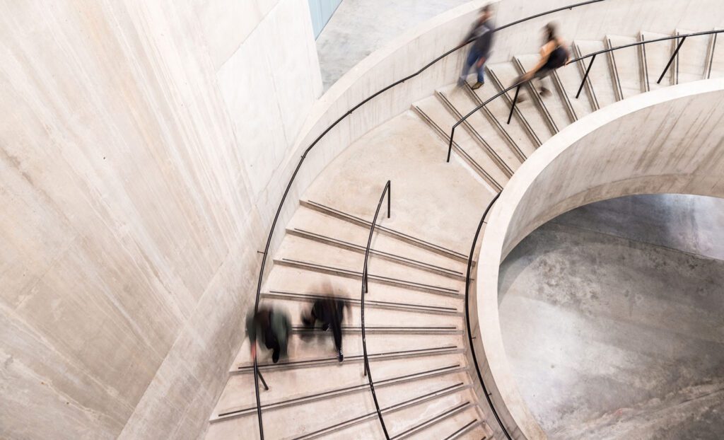 Color image depicting an abstract high angle view of a concrete spiral staircase. We can see the blurred motion of a group of people walking up and down the staircase, giving the impression that they are moving fast