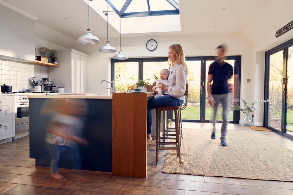 Busy Family Kitchen With Mother Working On Laptop And Holding Baby As Daughter Runs In Foreground