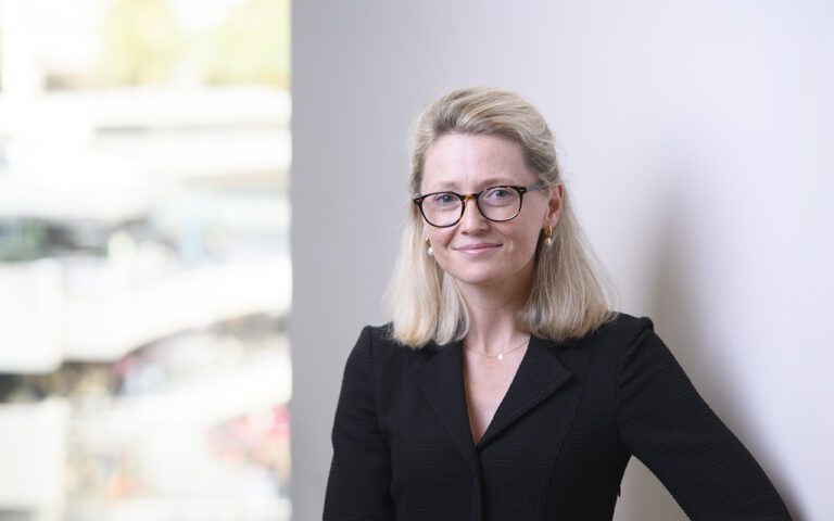 Molly Wills, a woman with blonde hair and glasses, wearing a black blazer, stands indoors near a window with a blurred background, smiling slightly.