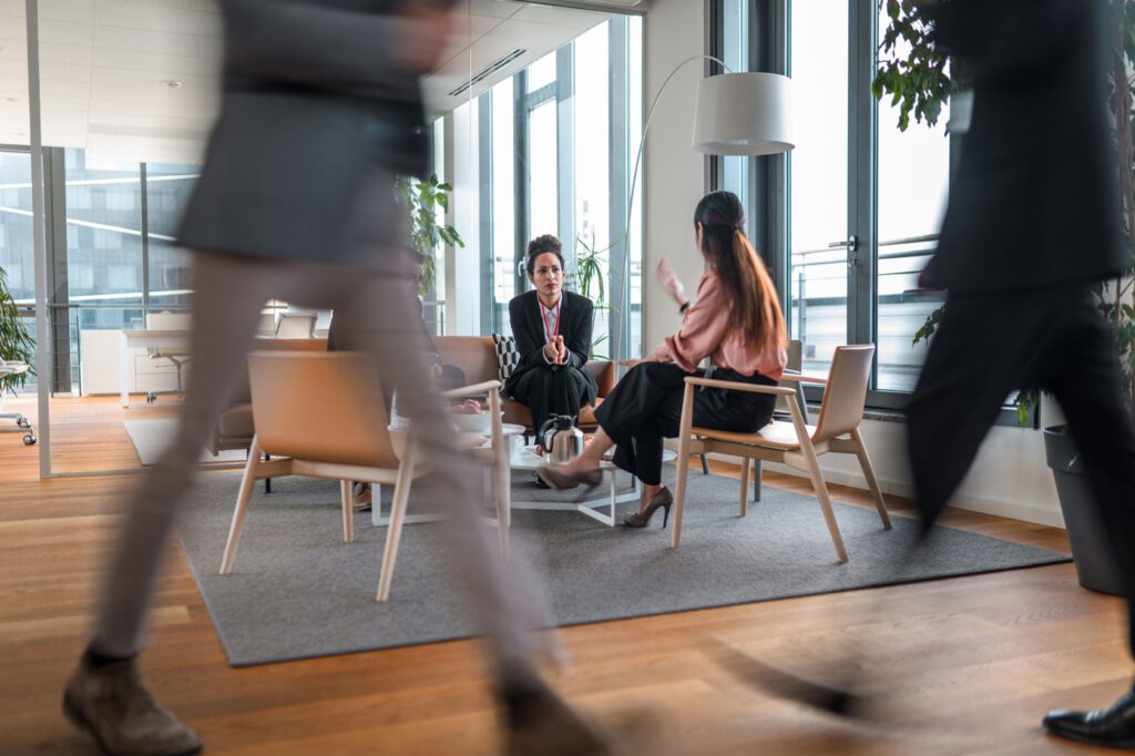 Two people sat in office speaking with blurry people walking infront