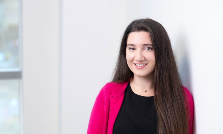 Niamh Meacock, a woman with long brown hair, wearing a black top and a bright pink cardigan, stands and smiles in front of a white wall with a window in the background.
