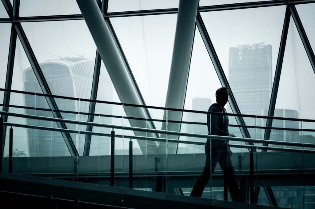 A single silhouetted businessman walking down the stairs of City Hall, a publically owned building which is accessible to the public in London, with the skyscrapers of the modern London skyline, including the walkie talkie building, in the background. The man is backlit and unrecognisable as he heads down the stairs.