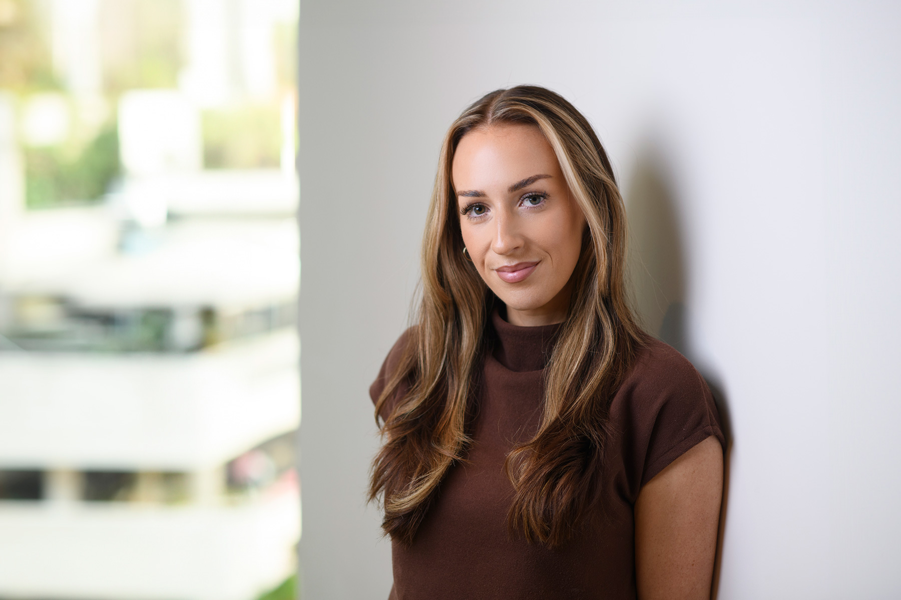 Emma Bryson smiling with long brown hair, wearing a brown short-sleeved top whilst leaning against a white wall.