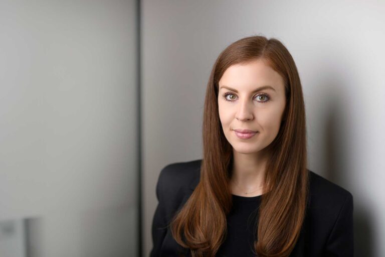 Moya Smith, a woman with long brown hair, wearing a black blazer, stands against a light-coloured wall and looks at the camera with a neutral expression.