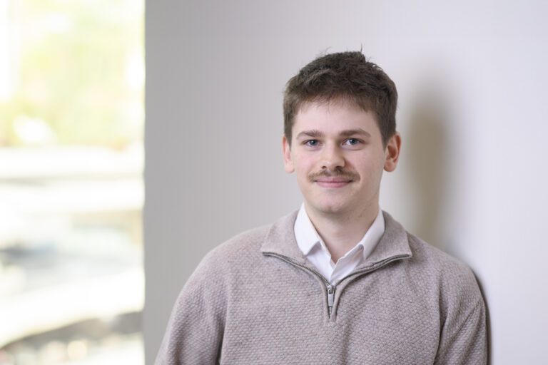 Harry Spice, a young man with a moustache, wears a beige quarter-zip jumper over a white collared shirt whilst leaning against a wall.