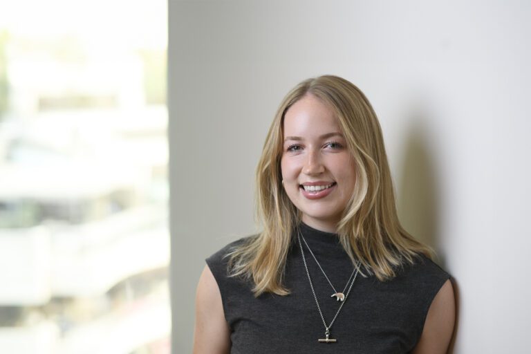 Meg Heaney is a smiling young woman with shoulder-length blonde hair, wearing a dark grey shirt and a silver pendant necklace.
