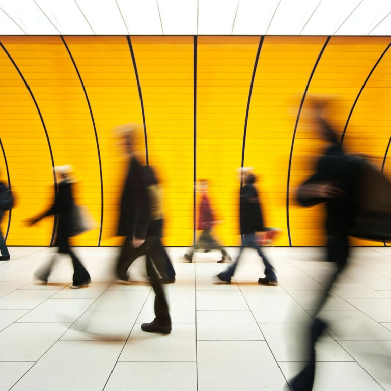 People walking briskly in front of a bright yellow, curved wall; motion blur emphasises their movement, while the background remains sharp and geometric.