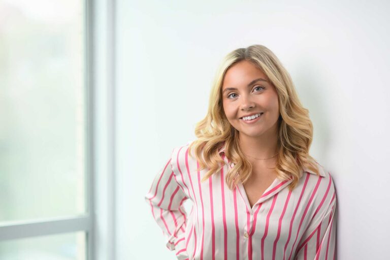 Ava Hill, a woman with long blonde hair wearing a pink and white striped shirt, stands indoors, smiling, with a window in the background.