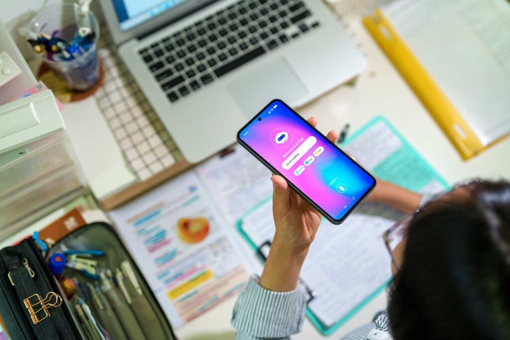 Student interacts with an AI chatbot on a smartphone while studying at a desk with a laptop, notes and stationery.