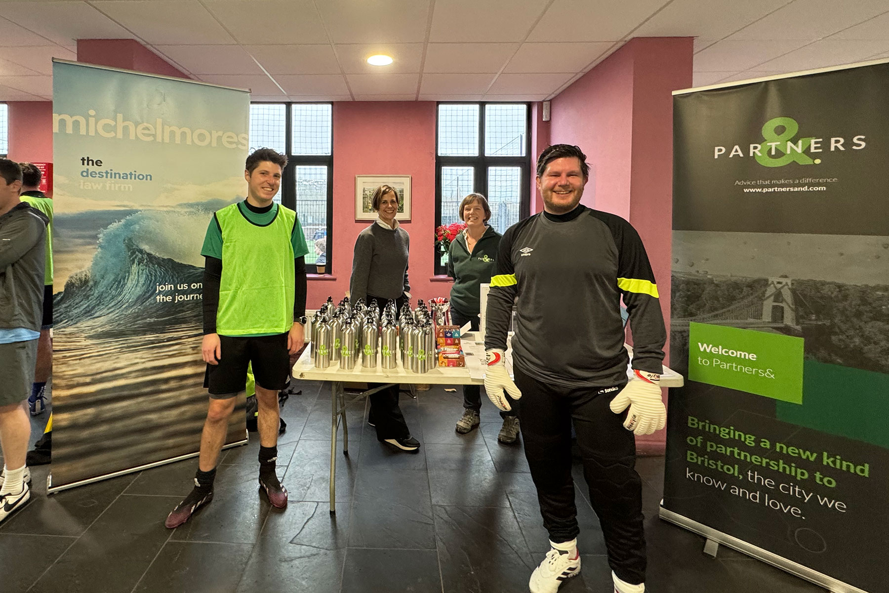 Four people stand indoors near tables with water bottles and banners for Michelmores law firm and Partners&. Two people wear sports kit and goalkeeper gloves; two others stand behind the table smiling.
