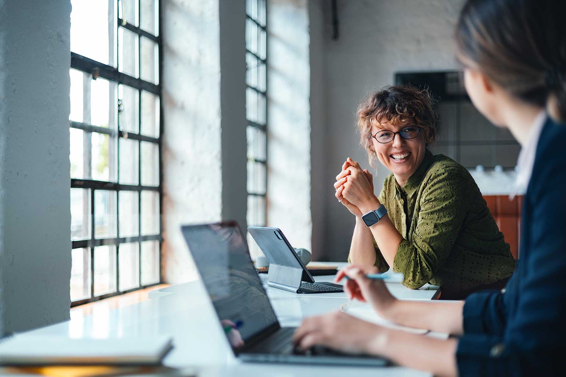 Two women collaborate in a bright coworking space, using laptops and a tablet