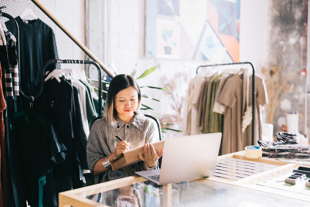emale clothing shop owner sits at her counter, writing on a box while her laptop is open, displaying shipping information