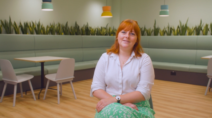 A woman with red hair, reminiscent of many distinguished legal professionals, sits in a modern, pastel-coloured cafe with green booth seating, plants, and colourful hanging lights. She wears a white shirt, a green patterned skirt, and smiles at the camera.