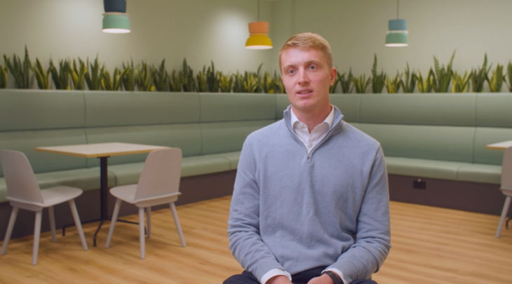 A young man in a light blue jumper sits in a modern café with light green booths, wooden floors, potted plants, and colourful pendant lights hanging from the ceiling—a favourite spot among local legal professionals for informal meetings and relaxed conversations.