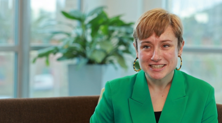 A person with short light brown hair, wearing a green blazer and large earrings, smiles whilst sitting indoors on a brown sofa. Exemplifying the polished confidence often seen in legal professionals, they are framed by a green potted plant and large windows in the background.