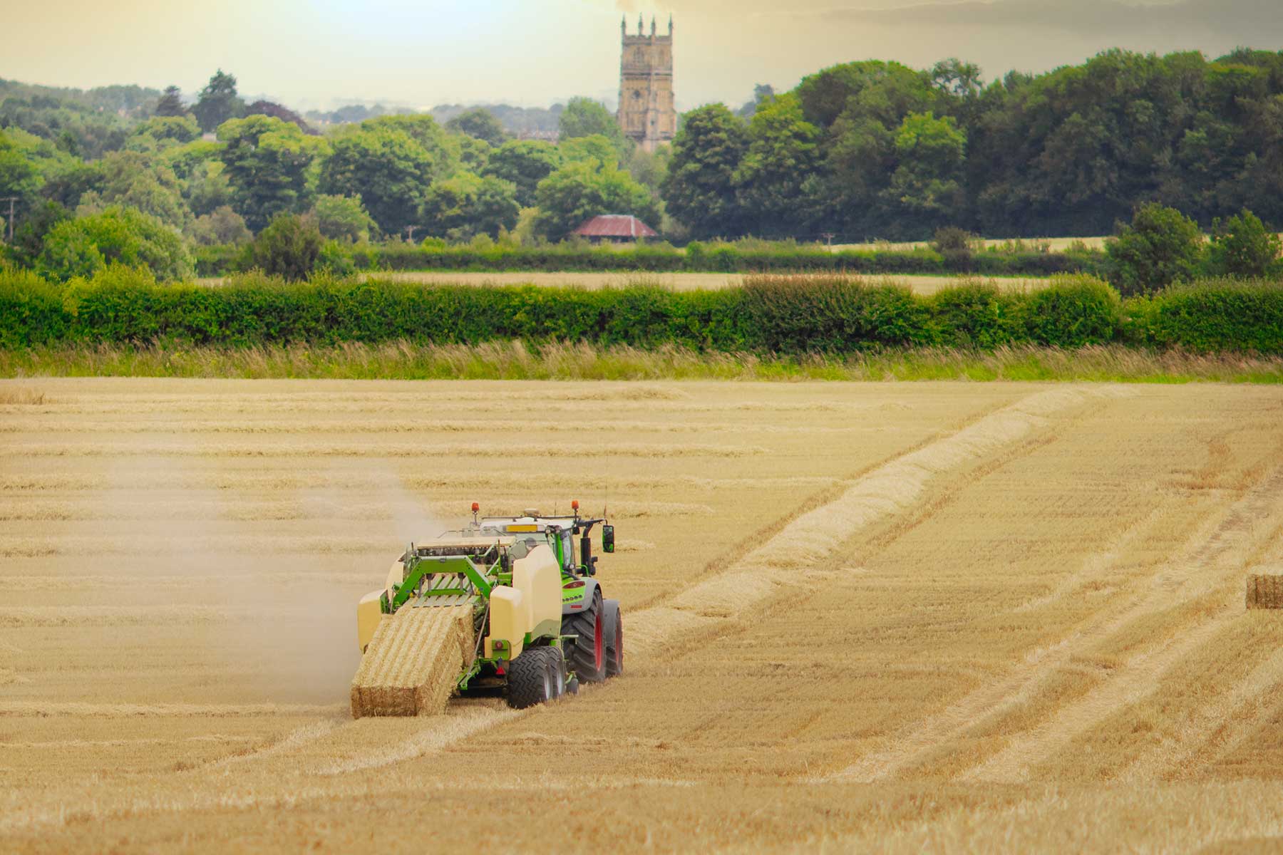 Straw or hay Baler machine pulled by a tractor, discharging a compacted straw bale from the rear in a field of recently harvested barley on the outskirts of Cirencester in the Cotswolds