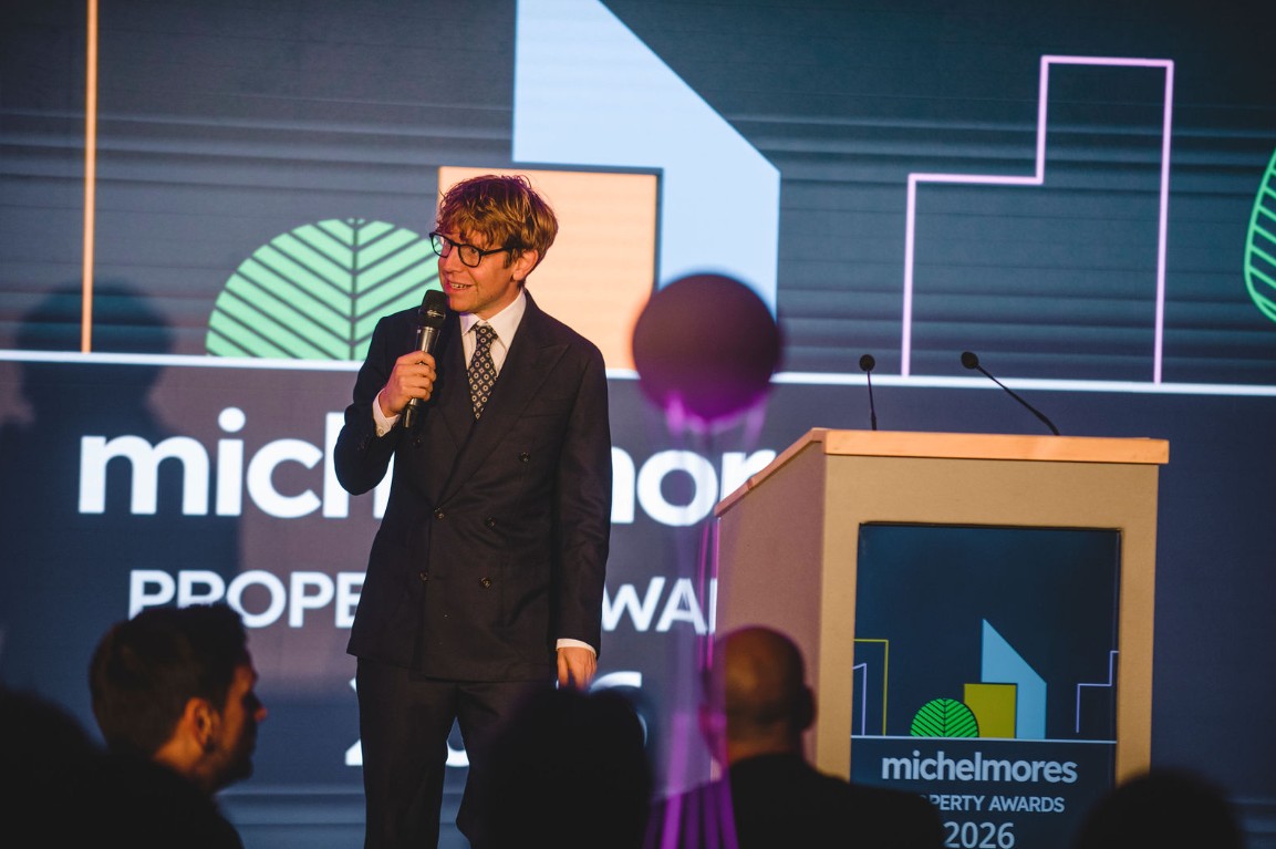 A man in a suit speaks into a microphone on stage at the Michelmores Property Awards 2026, standing beside a lectern with a lit backdrop and audience members visible in the foreground.