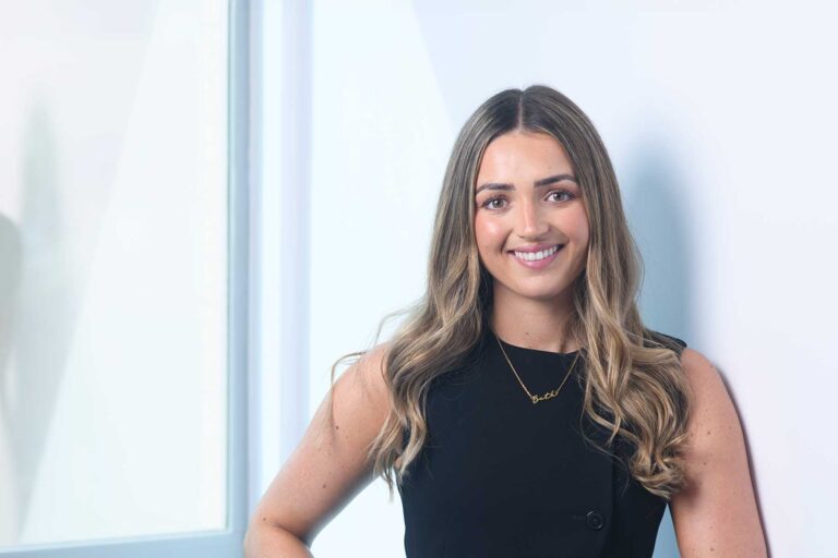 Bethany Lock, A woman with long, wavy hair wearing a sleeveless black top smiles whilst standing next to a window in a brightly lit room.