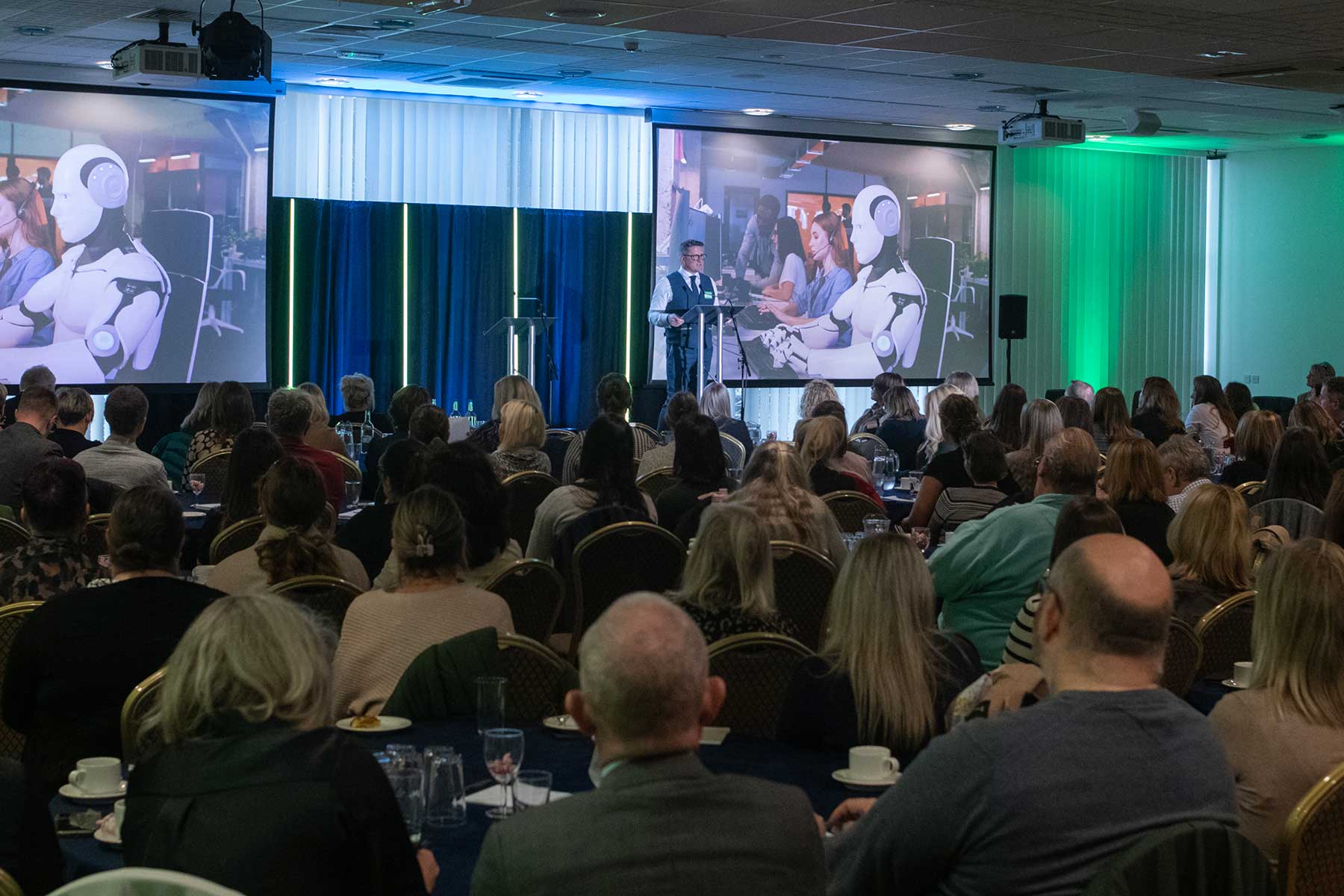 A speaker presents to an audience, with large screens behind showing images of humanoid robots interacting with people in an office setting.