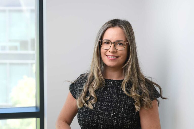 Image of Hannah Marshall. A woman with long, wavy blonde hair and glasses smiles, standing indoors near a window. She is wearing a short-sleeved, dark patterned top.
