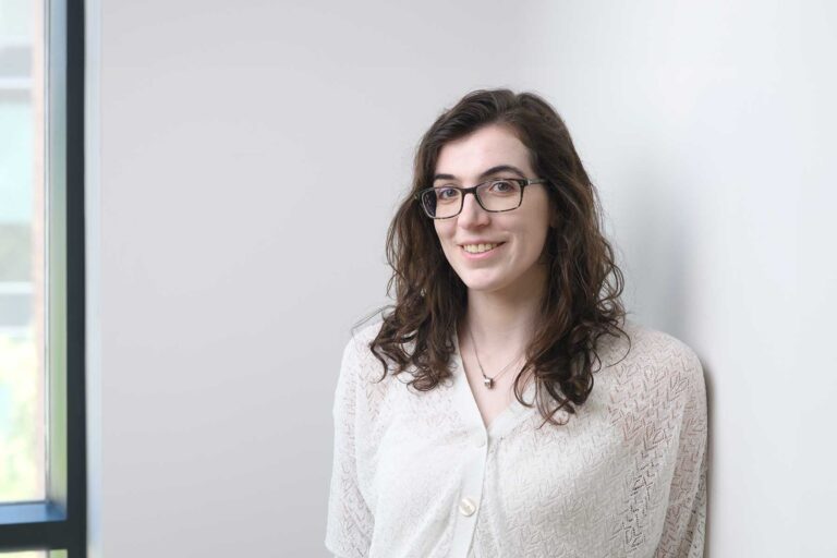 An image of Micol Flegenheimer. A woman with long brown hair and glasses, wearing a white patterned blouse, stands and smiles next to a white wall with a window nearby.