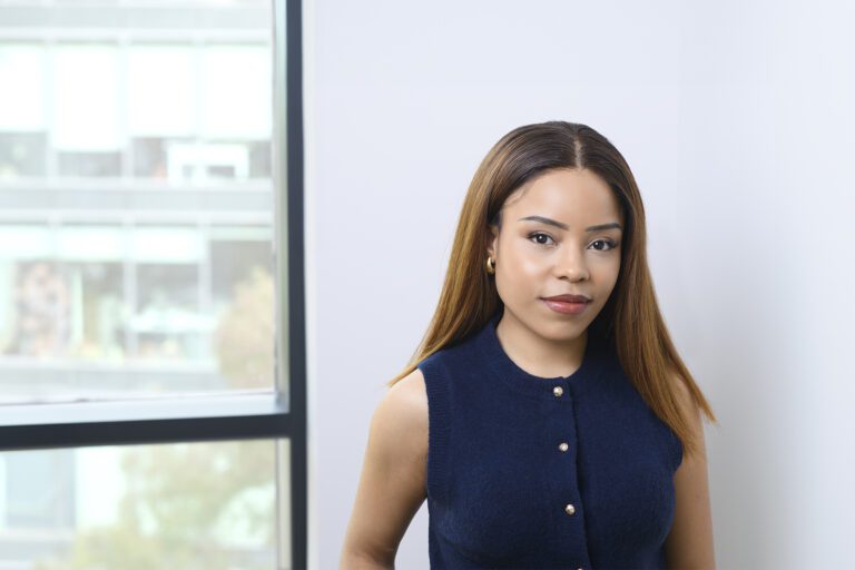 A woman with straight brown hair, wearing a sleeveless navy top, stands indoors near a window with an office block visible outside.