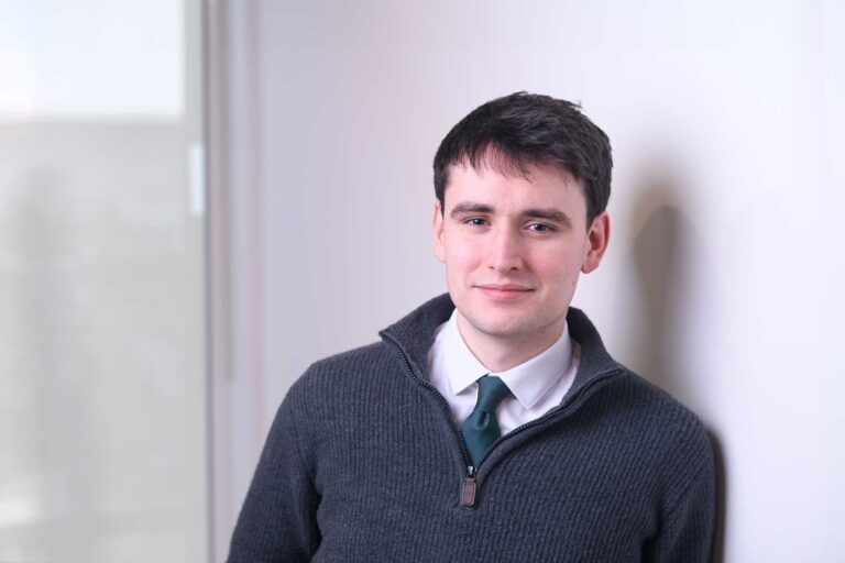 Elliot Thomas. A young man with short dark hair, wearing a dark jumper over a white shirt and green tie, stands indoors leaning against a white wall, looking at the camera with a slight smile.