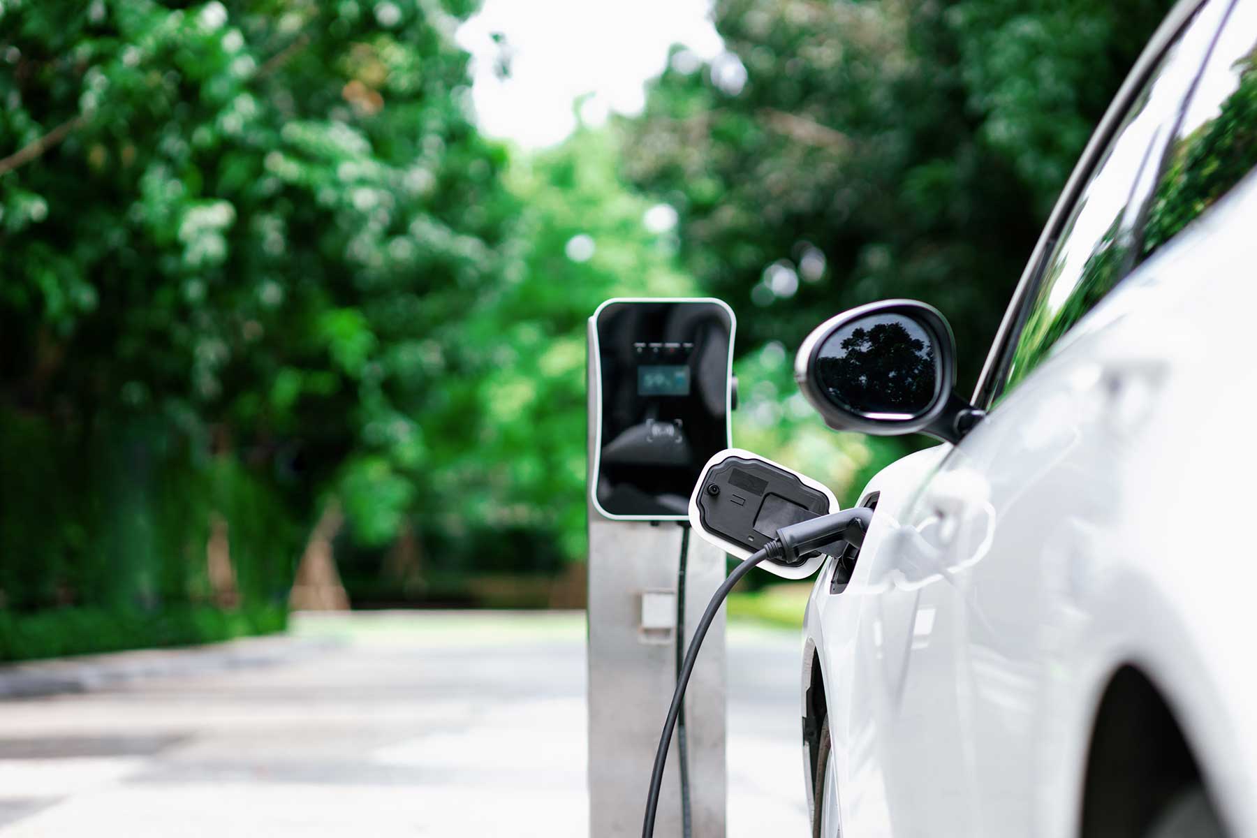 A white electric car is parked and plugged into a charging station outdoors, with green trees and a blurred background.