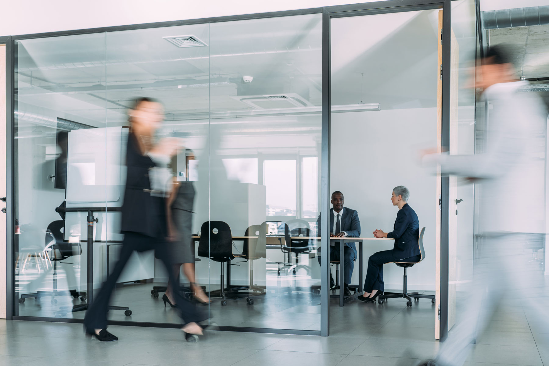 Two people sit and talk at a meeting table inside a glass-walled office, whilst blurred figures walk by outside, creating a sense of motion and busy office activity.