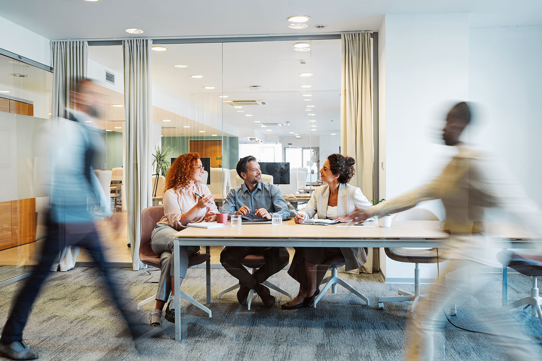 Three people sit at a conference table talking, while two blurred figures walk past them in a modern office with glass walls and bright lighting.