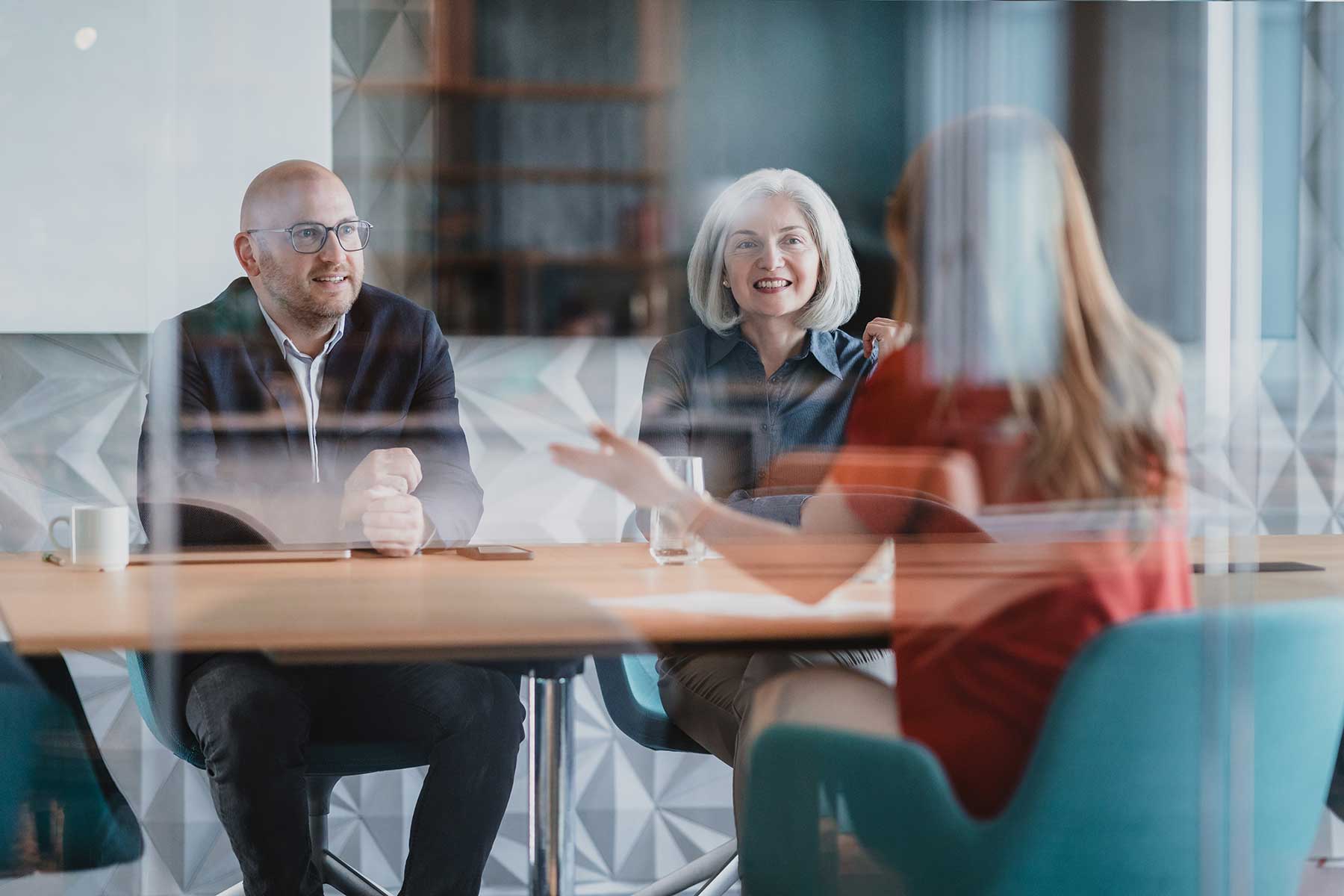 Three people sit at a conference table in a modern office, engaged in conversation. Two face the camera, one with grey hair and one bald, whilst a woman in red speaks with her back to the camera.