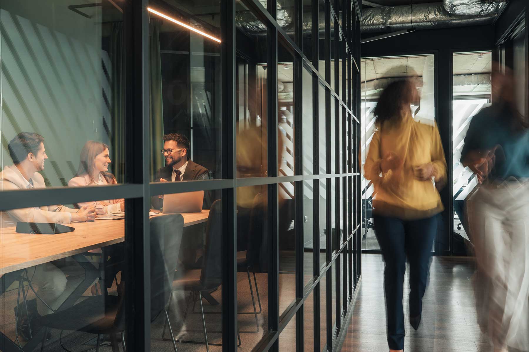Three people sit at a conference table in a glass-walled office having a discussion, whilst two others walk briskly down the corridor outside. The setting appears modern and professional.