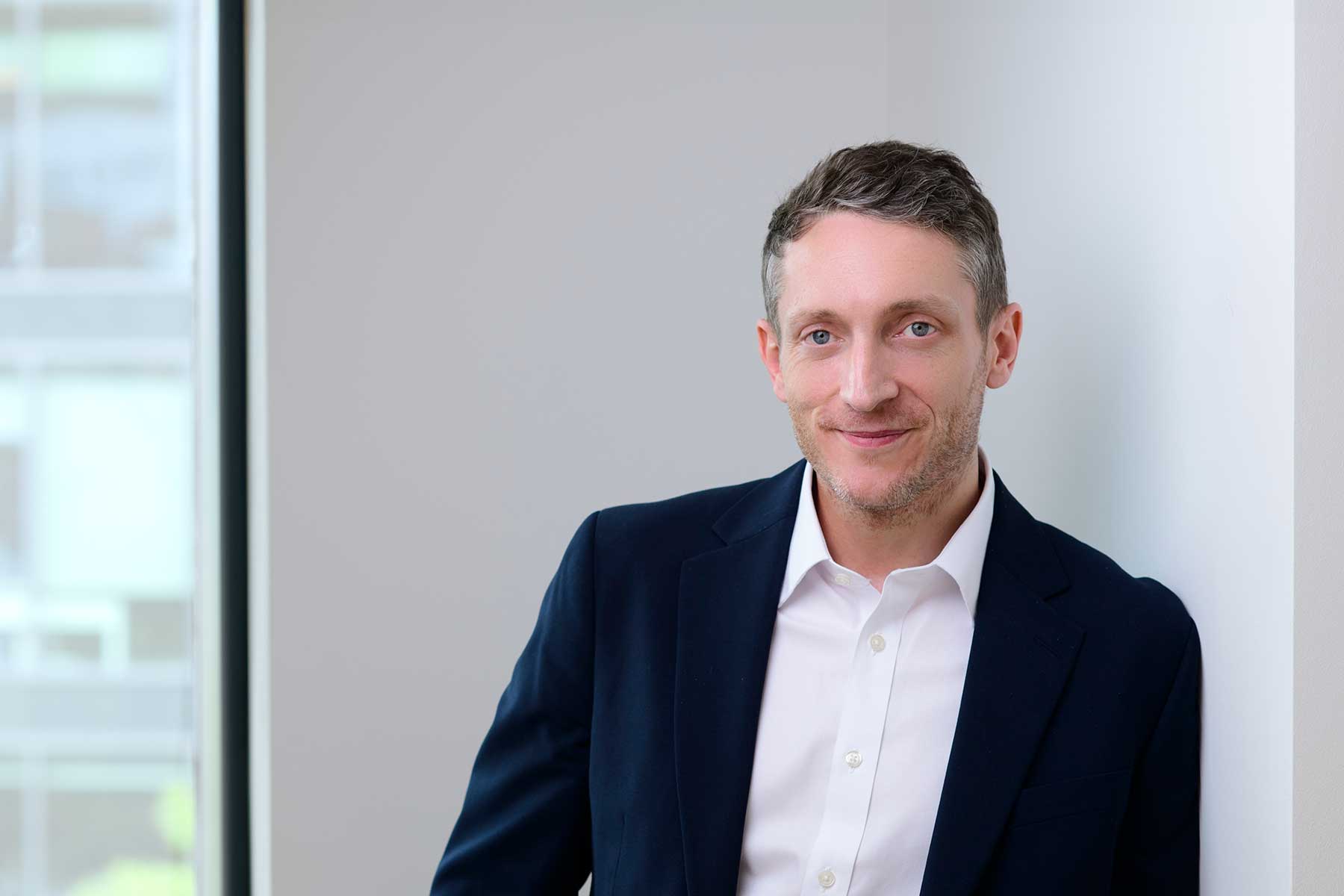 Sam Leyland. A man in a navy suit and white shirt leans against a white wall, smiling softly, with a bright window in the background.