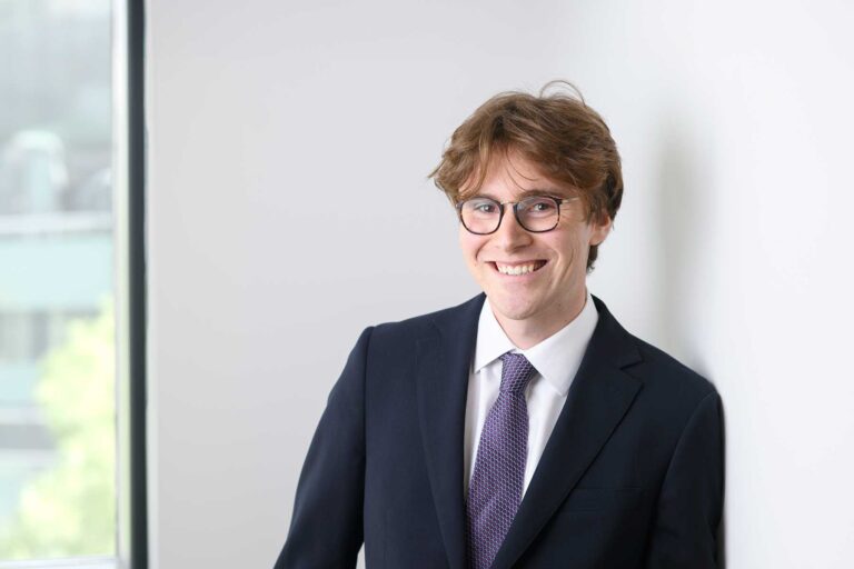 Ben Wilson. A young man with brown hair and glasses, wearing a dark suit, white shirt, and purple tie, smiling whilst standing near a window against a white wall.