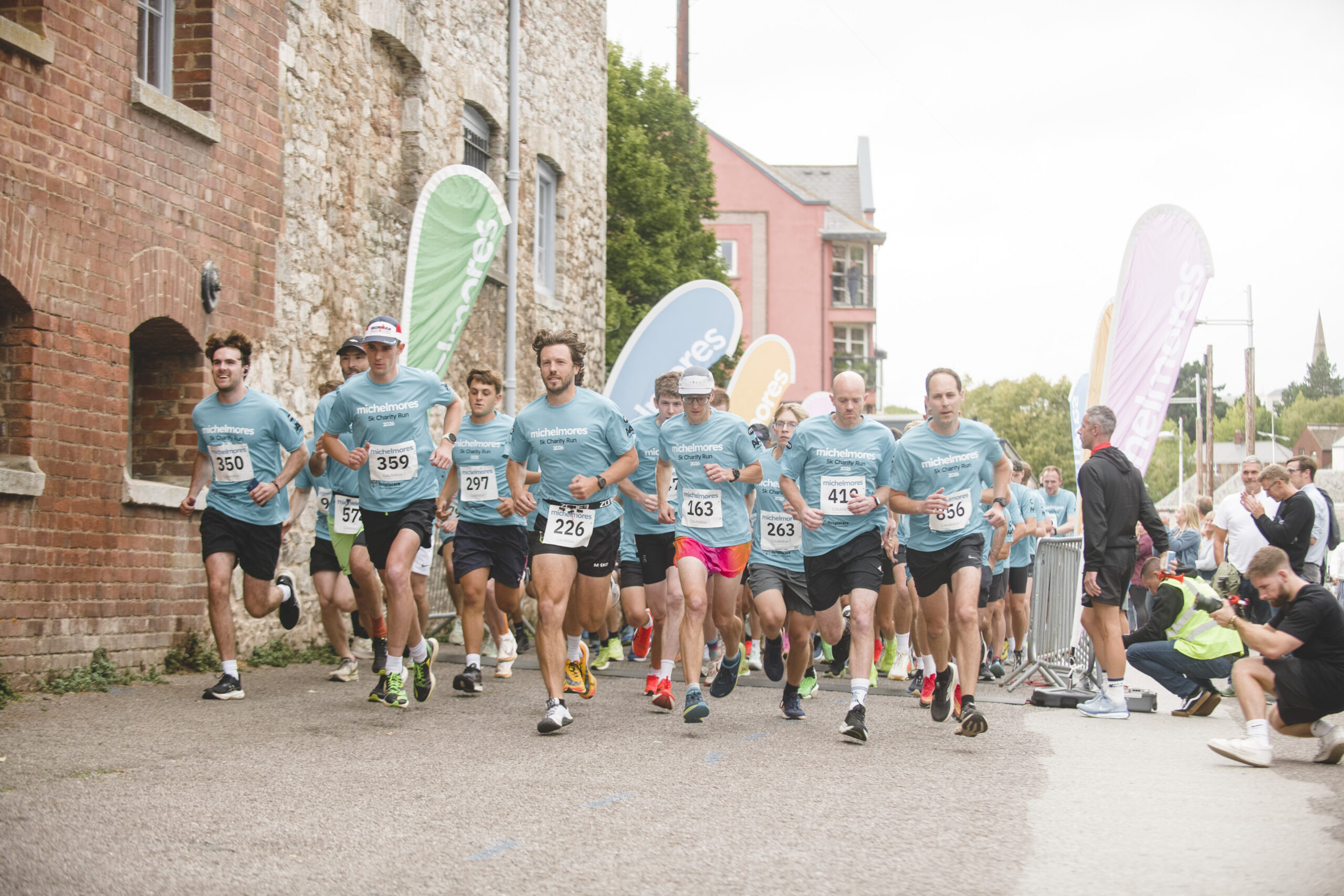 A group of runners wearing teal shirts and race numbers start a race near brick buildings, with colourful flags and spectators in the background.