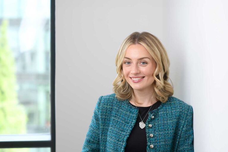 Ellie Knight. A woman with blonde hair in loose curls, wearing a teal textured jacket, smiles whilst standing indoors near a window with natural light.