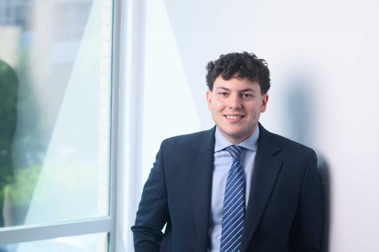 Emre Demir. A young man in a dark suit and striped tie is smiling whilst standing indoors near a large window with natural light coming in.